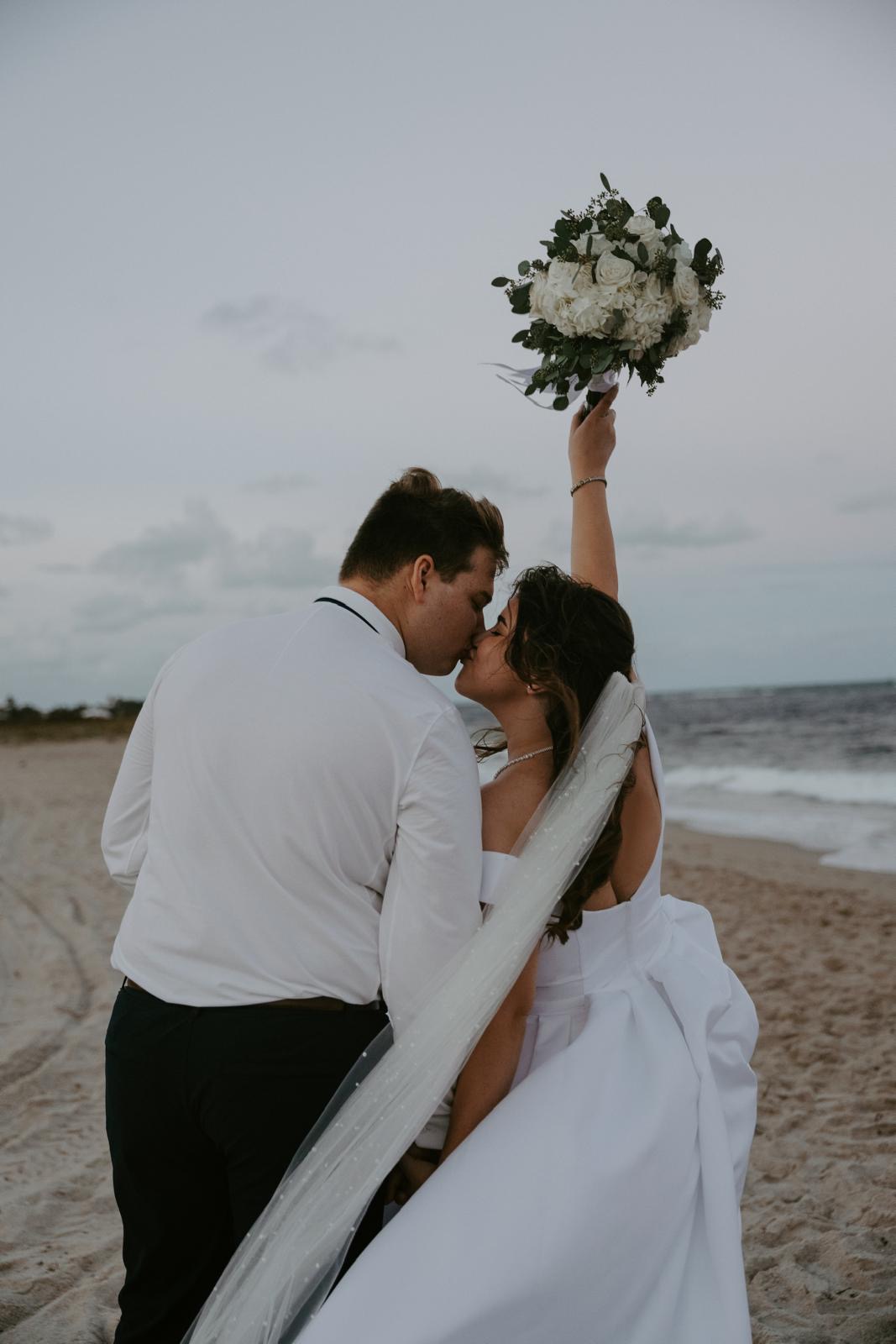 Newlyweds kissing as the bride raises her bouquet on a Florida beach