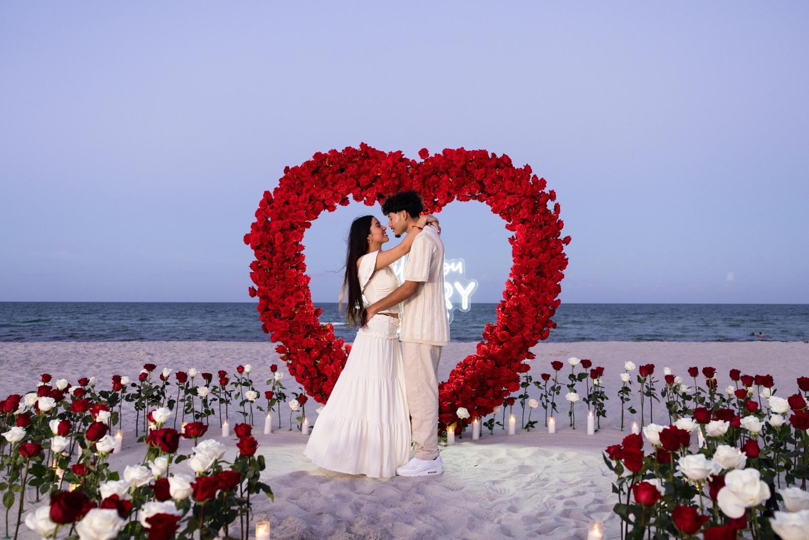 Couple embracing inside a red rose heart arch on Miami Beach at dusk