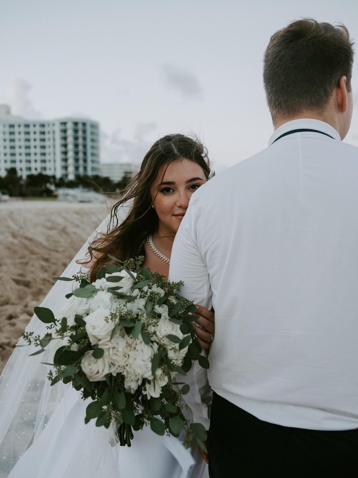 Bride with white rose bouquet glancing over the groom's shoulder on Miami Beach