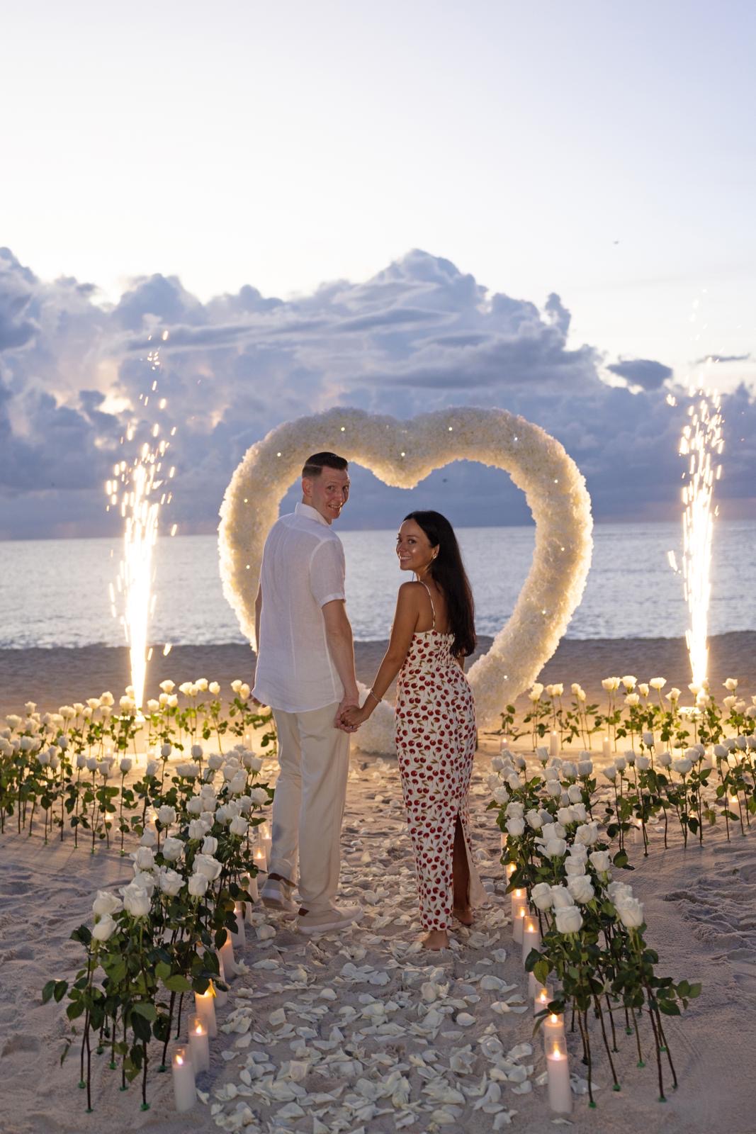 Couple holding hands before a white floral heart arch with sparklers at sunset on a South Florida beach